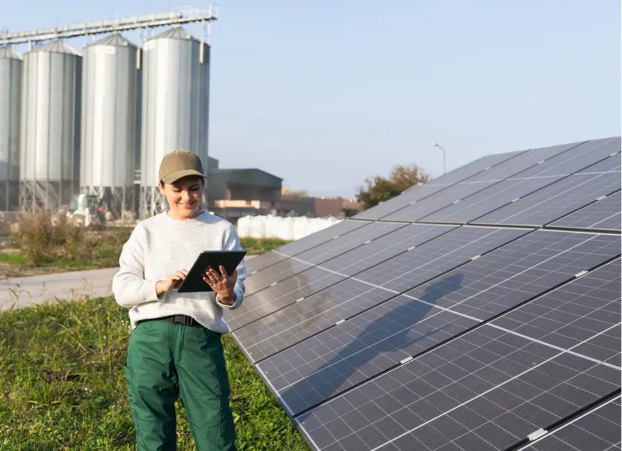 A woman wearing a beige cap, light gray sweater, and green work pants stands outdoors beside a large array of solar panels, holding and interacting with a digital tablet. She is smiling and appears focused on the screen. In the background, there are several large silver grain silos and industrial buildings, indicating a rural or agricultural setting integrating renewable energy technology. The sky is clear and blue.